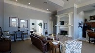 This single-floor communal space is designed to offer both comfort and style, making it an inviting gathering area for residents or guests. The main lounge area immediately draws attention with its soaring tray ceilings accented by crown molding, lending a sense of grandeur to the space. Neutral gray walls provide a sophisticated backdrop that complements the warm, rich tones of the leather seating and patterned accent chairs arranged around a central coffee table.

A prominent stone fireplace serves as the focal point of the lounge, framed by a stately white mantel supported by decorative columns. A mounted flat-screen television above the mantel adds a functional element to the elegant setting. Recessed lighting and ceiling fans contribute to the overall comfort and ambiance of the room.

Adjacent to the main lounge, an open doorway reveals a game room complete with a billiards table and bar-height seating, offering an additional layer of entertainment. Natural light filters in through windows visible in the background, brightening the connected spaces. The combination of classic architectural details, cozy furnishings, and versatile amenities makes this a well-appointed and functional communal environment.
