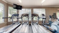 Located at 1500 Royal Gold Dr in Columbus, Ohio, this well-appointed fitness center offers a comprehensive workout environment on a single floor. The space is thoughtfully designed to accommodate a wide range of exercise routines, combining cardio and strength training areas within an open, accessible layout.

The cardio zone features multiple Life Fitness treadmills and an Echelon stationary bike, all positioned in front of large, white-shuttered windows that flood the room with natural light and provide pleasant views of the surrounding greenery. Wall-mounted flat-screen televisions enhance the cardio experience. Ceiling fans contribute to a comfortable atmosphere throughout.

The strength training area is generously equipped with a cable machine, Smith machine, weight benches, and a variety of free weights including kettlebells. Full-length mirrored walls line one side of the room, visually expanding the space and allowing users to monitor their form during workouts. The durable rubber flooring throughout provides both safety and functionality.

Additional amenities include a yoga mat station mounted on the wall, a wall clock, and ample open floor space for stretching or functional training. Crown molding and neutral warm-toned walls give the facility a polished, well-maintained appearance, making it a practical and inviting space for fitness enthusiasts.