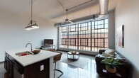 This single-floor unit showcases a compelling industrial aesthetic rooted in its conversion from a warehouse or factory-style building. The living area serves as the centerpiece of the space, anchored by a dramatic floor-to-ceiling multi-pane steel window wall that spans nearly the entire exterior facade, flooding the interior with natural light and offering views of the surrounding brick architecture. Exposed metallic ductwork runs overhead against a whitewashed wood-panel ceiling, reinforcing the industrial character, while polished concrete floors flow consistently throughout the main living areas.

The open-concept layout connects the living room seamlessly to the kitchen, which features rich dark wood cabinetry, white countertops, a stainless steel refrigerator, and a peninsula with bar seating. A hallway with textured plaster walls leads to the private areas of the unit. The bedroom offers a quiet retreat with polished terrazzo-style flooring and recessed ceiling lighting. Two bathrooms are present, each with a tub-shower combination and dark wood vanities with white countertops. A dedicated in-unit laundry room with a full-size washer and dryer completes the layout, adding practical convenience to this characterful industrial-style space.