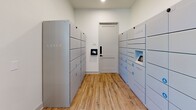 Located at 225 Rhyne Rd in Charlotte, NC, this single-floor mail and package management facility is thoughtfully designed to serve a residential community with efficiency and a clean, contemporary aesthetic. The space is organized into distinct functional zones, each purpose-built to handle resident mail and parcel deliveries seamlessly.

The interior features wood-look tile flooring throughout, lending a warm yet durable finish that ties the space together. Crisp white walls and recessed lighting create a bright, welcoming environment, while pendant light fixtures add a polished design touch above key areas.

A dedicated package locker room houses multiple Luxer One smart locker units lining both walls, offering secure, automated package pickup for residents of varying parcel sizes. The room is accessed through a keyed entry door and includes a digital touchscreen interface for streamlined locker management.

Adjacent to the package area, a generously sized mailroom corridor accommodates an extensive bank of individual resident mailboxes mounted along central dividing structures. Tall windows near the entry allow natural light to filter in, enhancing visibility and openness. The layout provides comfortable circulation throughout, and the overall design reflects a modern, functional approach to community mail services.