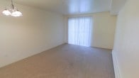 This single-floor space presents a clean and understated design, built around a neutral color palette that creates a calm and inviting atmosphere. The interior is finished with wall-to-wall carpeting in a warm beige tone, providing a soft and comfortable underfoot feel that complements the light-colored walls throughout. Natural light is a defining feature of the space, with a large window fitted with vertical blinds that allow for flexible light control while brightening the room with an airy, open feel. The smooth, light-toned ceiling enhances the sense of openness within the space. The overall layout is open and unencumbered, offering flexibility for a variety of furniture arrangements and functional uses. The simple, unadorned design ensures the space serves as a versatile canvas, well-suited to accommodate different styles and preferences. The combination of soft carpeting, neutral walls, and generous natural light gives the space a welcoming and comfortable ambiance throughout.
