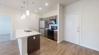 Located at 17744 Wayforest Dr in Houston, Texas, this single-story residence offers a thoughtfully designed, open-concept layout with modern finishes throughout. The kitchen serves as the centerpiece of the living area, featuring a large island with a built-in sink and white countertops, dark cabinetry, a subway tile backsplash, and stainless steel appliances including a full-size refrigerator, electric range, microwave, and dishwasher. Pendant lighting hangs above the island, complementing the recessed ceiling lights that brighten the space. The kitchen flows seamlessly into the living area, which is outfitted with wood-look flooring and a ceiling fan, creating a comfortable and cohesive common space. A front door with a sidelight window allows natural light to enter the living area. The bedrooms feature a mix of wood-look and carpeted flooring, with ceiling fans and access to natural light through window blinds offering views of the surrounding greenery. The bathrooms are finished with light tile flooring; one includes a soaking tub with a white subway tile surround, while another features dark cabinetry, a white countertop vanity, and a wall-mounted grab bar. Throughout the home, neutral tones and clean lines create a bright, modern ambiance.