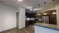 This single-floor space showcases a thoughtfully designed open-concept layout that seamlessly integrates the kitchen and living areas. The kitchen is a standout feature, appointed with rich dark wood cabinetry that provides ample storage and a bold visual contrast against the classic white subway tile backsplash. Stainless steel appliances, including a refrigerator, electric range, and built-in microwave, offer both functionality and a polished, contemporary aesthetic. A granite-topped island anchors the kitchen, illuminated by sleek pendant lights that add warmth and character to the space. Track lighting overhead ensures the entire kitchen area is well-lit. The flooring throughout is durable wood-look planking in a warm tone, contributing to a cohesive and inviting atmosphere. Clean white walls and smooth ceilings keep the interior feeling bright and open. A gray-trimmed interior door visible in the space adds a subtle modern touch to the overall design. The combination of upscale finishes and practical layout makes this a highly functional and visually appealing living space.