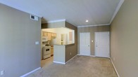 This single-floor residence offers a practical and comfortable open-concept layout designed to maximize both function and flow. The living area features plush wall-to-wall carpeting and is bathed in natural and recessed lighting, creating a warm and inviting atmosphere. Crown molding and vaulted ceilings add an architectural touch that elevates the overall feel of the space.

The kitchen is positioned adjacent to the living area and is accessible via a pass-through counter with a raised breakfast bar, making it ideal for casual dining or entertaining. The kitchen is equipped with white appliances, including a range and microwave, and features light wood cabinetry that provides ample storage. Under-cabinet lighting enhances visibility and adds a subtle warmth to the cooking space.

Neutral taupe-toned walls run throughout the unit, providing a cohesive and versatile backdrop that complements a variety of décor styles. Two white paneled doors are visible from the living area, likely leading to additional rooms or storage, contributing to the unit's functional layout. Recessed lighting in the ceiling and a centrally placed HVAC vent highlight the thoughtful construction of this well-maintained, move-in-ready space.