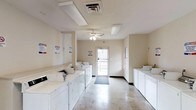 This single-floor laundry facility is designed for convenient, communal use. The space features rows of white washing machines and dryers lining both sides of the room, providing ample capacity for residents. The interior is finished with light-toned walls and a polished concrete floor that is easy to maintain and clean. Overhead, a ceiling fan complements the recessed fluorescent lighting to keep the space well-ventilated and brightly lit. A central glass-panel door at the far end of the room allows natural light to filter in while providing direct exterior access. A window along the side wall further enhances the natural brightness of the space. Informational and instructional signage is posted throughout the facility to assist users with equipment operation and facility guidelines. The layout is open and uncluttered, allowing for easy movement between machines. Overall, the facility offers a clean, practical, and well-organized environment suited to everyday laundry needs.