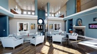 Located in Dallas, this single-floor space presents a grand, open lobby designed to impress. The most commanding feature is the dramatic double-height ceiling, which creates an expansive, airy atmosphere and is complemented by rows of clerestory windows that flood the interior with abundant natural light. Towering teal-painted columns and accent walls provide a bold yet refined color palette against crisp white trim and neutral ceiling tones.

The polished dark hardwood floors reflect the recessed lighting above, enhancing the sense of depth and luxury throughout the space. Multiple seating arrangements featuring contemporary white upholstered sofas, armchairs, and low-profile coffee tables are thoughtfully positioned to encourage both intimate conversation and open gathering.

Near the entrance, a distinctive circular architectural element frames the entryway, while colorful stained-glass-style wall art panels add an artistic focal point. To one side, a cozy lounge nook includes a fireplace, a media display, and additional seating, offering a more relaxed retreat within the larger open plan. The overall design balances modern elegance with a warm, inviting ambiance, making this Dallas space both visually dynamic and functionally versatile for social interaction.