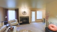 This single-floor space centers around a welcoming living area finished in warm neutral tones throughout. The room features a wood-burning fireplace framed by dark granite or slate surround and hearth, serving as the focal point of the space. Above the fireplace, a decorative sunburst mirror adds a stylish touch to the otherwise understated wall. Track lighting mounted on the ceiling provides adjustable illumination, complementing the natural light that filters in through multiple windows dressed with horizontal blinds and accented by rich brown curtain panels. A glass-paneled exterior door allows additional daylight into the room while offering access to the outdoors. The carpeted flooring in soft beige tones contributes to the room's comfortable, lived-in feel, and a cowhide-style area rug adds a subtle textural contrast near the seating area. White upholstered furniture creates a clean, contemporary contrast against the warmer wall tones. A small dining or seating area is partially visible near the rear window, suggesting a functional layout that transitions naturally from the main living space. Overall, the space presents a relaxed and inviting atmosphere with practical design elements throughout.