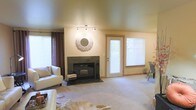 This single-floor space centers around a welcoming living area finished in warm neutral tones throughout. The room features a wood-burning fireplace framed by dark granite or slate surround and hearth, serving as the focal point of the space. Above the fireplace, a decorative sunburst mirror adds a stylish touch to the otherwise understated wall. Track lighting mounted on the ceiling provides adjustable illumination, complementing the natural light that filters in through multiple windows dressed with horizontal blinds and accented by rich brown curtain panels. A glass-paneled exterior door allows additional daylight into the room while offering access to the outdoors. The carpeted flooring in soft beige tones contributes to the room's comfortable, lived-in feel, and a cowhide-style area rug adds a subtle textural contrast near the seating area. White upholstered furniture creates a clean, contemporary contrast against the warmer wall tones. A small dining or seating area is partially visible near the rear window, suggesting a functional layout that transitions naturally from the main living space. Overall, the space presents a relaxed and inviting atmosphere with practical design elements throughout.