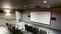 This dedicated screening room is designed to deliver a true theater experience within a private setting. The space features tiered, stadium-style seating arranged across multiple rows, ensuring unobstructed sightlines to the large projection screen mounted on the front wall. The seating consists of dark upholstered chairs equipped with built-in cup holders, prioritizing comfort during extended viewing sessions. A metal railing runs along the aisle, reinforcing the tiered layout and providing safe navigation between rows. The room is finished with neutral-toned walls and a dark ceiling, both of which help minimize light reflection and enhance on-screen image quality. Recessed ceiling lights provide ambient illumination when the room is not in use, while HVAC vents ensure climate control throughout. A wall-mounted speaker is positioned to one side of the screen, contributing to the room's audio setup. A small window on the side wall introduces a subtle connection to the exterior. The overall design is functional and focused, creating an environment well-suited for film screenings, presentations, or private events.