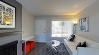 This single-floor space offers a clean, modern aesthetic with a thoughtfully arranged interior. The living area serves as the centerpiece of the layout, featuring smooth grey walls and wood-style flooring that lend a contemporary, cohesive feel throughout. A large front-facing window fitted with vertical blinds allows natural light to fill the room, creating a bright and welcoming atmosphere. The living space is furnished with a white sofa accented by decorative pillows, a round glass coffee table with a sleek chrome base, and a plush grey area rug that anchors the seating arrangement. Vibrant red ottomans provide a bold pop of color alongside a minimalist white desk, adding both function and personality to the space. A floor lamp in the corner contributes warm ambient lighting. The entry door is positioned directly within the living area, contributing to the open and accessible flow of the floor plan. The overall ambiance is modern and efficiently utilized, making the most of the available space with carefully selected furnishings that balance comfort and style. Natural light and neutral wall tones keep the interior feeling open and airy throughout the day.