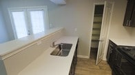This single-floor space showcases a thoughtfully designed kitchen area that balances functionality with a clean, contemporary aesthetic. The kitchen features smooth white countertops that extend generously along the wall, providing ample prep and workspace. A stainless steel undermount sink sits beneath a window fitted with white plantation shutters, allowing natural light to filter into the space while maintaining privacy. The dark-toned cabinetry below the counters offers a subtle contrast to the bright surfaces above. Wood-look flooring runs throughout, adding warmth and a sense of continuity to the overall design. A notable feature of this kitchen is the adjacent walk-in pantry, accessible through a paneled door. The pantry is equipped with multiple built-in shelves, offering generous and well-organized storage space for dry goods and kitchen essentials. The walls throughout are finished in a neutral light gray tone, contributing to a calm and cohesive atmosphere. Overall, the space presents a practical and well-appointed layout that prioritizes storage, natural light, and a modern design sensibility.