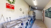 Located at 1540 Mosaic Way in Stockton, California, this shared laundry facility is a compact yet efficiently organized single-room space designed to serve the needs of its users. The room features light-toned hardwood-style flooring throughout, clean neutral walls, and a combination of overhead fluorescent lighting and a skylight that together provide ample brightness.

Along one wall, a row of LG front-load washing machines is neatly arranged side by side, each equipped with individual water hookups and card-based payment systems. On the opposite end of the room, stacked LG washer and dryer units maximize the use of vertical space, with industrial-style exhaust ducts routed along the ceiling for ventilation. A wall-mounted folding table provides a convenient surface for sorting and folding laundry.

The room includes thoughtful decorative touches such as a floral wreath and wall signage, adding a welcoming feel to an otherwise functional space. Posted notices and laundry rules are clearly displayed for users. A large sliding glass door along one side floods the room with natural light and offers a view of the surrounding greenery, creating an open and airy atmosphere. The space is well-maintained and organized, making it a practical and pleasant shared laundry facility.