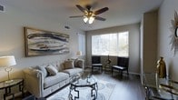 This single-story space showcases a thoughtfully designed living room that combines comfort and contemporary style. The room is anchored by a plush, tufted sofa dressed in neutral tones and complemented by a pair of sleek, dark leather accent chairs arranged near a large window. A geometric glass-top coffee table sits atop a patterned area rug, adding a modern focal point to the seating arrangement.

The flooring throughout is hardwood-style, providing a clean and cohesive look that enhances the room's overall aesthetic. A five-blade ceiling fan with an integrated light fixture hangs overhead, contributing both function and a classic decorative touch. The walls are finished in a soft, neutral tone, creating a calm and versatile backdrop.

Natural light flows generously into the space through a wide window fitted with vertical blinds, offering both brightness and privacy control. Outdoor greenery is visible through the window, adding a pleasant connection to the exterior environment. A large abstract artwork adorns one wall, lending a creative and refined character to the space. Overall, the living area presents a balanced blend of elegance and practicality.