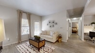 This single-floor residence offers a thoughtfully designed layout comprising three bedrooms, one bathroom, a combined living and dining area, a kitchen, a closet, and a connecting hallway. The living room greets visitors with light wood-style flooring, neutral gray walls, and multiple windows dressed in patterned curtains that allow generous natural light to fill the space. A front entry area provides a welcoming transition into the home. The kitchen is efficiently arranged with white cabinetry, light quartz-style countertops, a classic subway tile backsplash, stainless steel appliances including a range, microwave, and dishwasher, and a double-basin sink. Adjacent to the kitchen, the dining area accommodates a round dining table beneath a flush ceiling light, with hardwood-style flooring continuing the cohesive aesthetic. The bedrooms feature plush carpet flooring and ceiling fans, with the primary bedroom offering a comfortable, well-proportioned retreat. The shared full bathroom includes a tub-shower combination, vanity with white cabinetry, and light tile flooring. Throughout the residence, a consistent palette of soft whites and warm grays creates a clean, modern ambiance well-suited for everyday living.