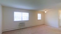 This single-floor space offers a clean, functional layout with a neutral and understated aesthetic. The main living area is finished with light beige carpet that extends throughout, creating a soft, cohesive feel underfoot. The room is well-lit by natural light streaming in through three white-framed windows fitted with horizontal blinds, two positioned side by side along one wall and a third on an adjacent wall, providing cross ventilation potential and a bright atmosphere even without artificial lighting. A ceiling fan is centrally mounted, offering both air circulation and supplemental lighting. Climate control is well-covered, with a wall-mounted air conditioning unit situated beneath the double windows and baseboard heating running along the perimeter walls, ensuring year-round comfort. The walls are painted in a clean, off-white tone that complements the light flooring and contributes to the open, airy feel of the space. The overall layout is straightforward and versatile, suitable for a range of furnishing arrangements. The space presents as move-in ready with practical amenities already in place.
