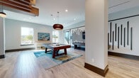 Located in Jacksonville, FL, this contemporary game room is thoughtfully designed to offer a dynamic social and recreational experience. The space features light wood-look flooring throughout, complemented by crisp white walls accented with a striking vertical slat wall detail that adds architectural interest and visual depth.

At the heart of the room sits a substantial dark wood pool table, anchored by a vibrant blue abstract area rug and illuminated by a sculptural copper pendant light above. Adjacent to this, a foosball table provides additional entertainment in an open corridor-style layout that keeps the space feeling airy and connected.

A cozy lounge nook offers built-in wraparound bench seating upholstered in teal and gray tones, accented with colorful throw pillows and centered around a unique wood-slice coffee table beneath a woven pendant light. Nearby, a high counter with wooden bar stools provides a casual perch beneath large windows that draw in abundant natural light.

The space also includes a well-appointed coffee and refreshment bar, complete with a coffee machine, mini refrigerator, and sink, finished with warm wood cabinetry and pendant lighting. Contemporary artwork adorns the walls throughout, tying together the coastal-inspired color palette of blues, teals, and warm neutrals for a cohesive, inviting ambiance.