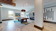 Located in Jacksonville, FL, this contemporary game room is thoughtfully designed to offer a dynamic social and recreational experience. The space features light wood-look flooring throughout, complemented by crisp white walls accented with a striking vertical slat wall detail that adds architectural interest and visual depth.

At the heart of the room sits a substantial dark wood pool table, anchored by a vibrant blue abstract area rug and illuminated by a sculptural copper pendant light above. Adjacent to this, a foosball table provides additional entertainment in an open corridor-style layout that keeps the space feeling airy and connected.

A cozy lounge nook offers built-in wraparound bench seating upholstered in teal and gray tones, accented with colorful throw pillows and centered around a unique wood-slice coffee table beneath a woven pendant light. Nearby, a high counter with wooden bar stools provides a casual perch beneath large windows that draw in abundant natural light.

The space also includes a well-appointed coffee and refreshment bar, complete with a coffee machine, mini refrigerator, and sink, finished with warm wood cabinetry and pendant lighting. Contemporary artwork adorns the walls throughout, tying together the coastal-inspired color palette of blues, teals, and warm neutrals for a cohesive, inviting ambiance.