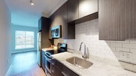 This well-appointed single-floor residence offers a thoughtfully designed layout that balances style and functionality across its key living spaces. The kitchen is a standout feature, showcasing dark espresso-toned cabinetry that extends to the ceiling, complemented by granite-look countertops and a classic white subway tile backsplash. Stainless steel appliances — including a refrigerator, electric range, over-the-range microwave, and dishwasher — provide a complete, modern cooking environment. Hardwood-style flooring runs throughout the main living and kitchen areas, contributing to a warm, cohesive aesthetic. The open living area benefits from generous natural light courtesy of large windows fitted with horizontal blinds, offering views of the surrounding urban landscape. The bedroom is carpeted for comfort and includes a sliding-door closet with built-in shelving and hanging space, providing ample storage. The bathroom features light-toned tile flooring, a dark wood vanity with a clean white countertop, and neutral wall tones that create a calm, understated atmosphere. Overall, the space presents a clean, contemporary design with consistent finishes throughout, making it a practical and visually cohesive living environment.