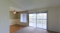 This single-floor space showcases a bright and functional living area designed to maximize natural light and comfort. The main living space features a combination of carpet and hardwood-style flooring, creating a clear visual distinction between different zones within the room. A textured ceiling is complemented by a ceiling fan with an integrated light fixture, providing both ventilation and overhead lighting.

Two-tone wall treatment adds a subtle design element, with the lower portion featuring a lighter finish that transitions to a muted green-grey tone above, creating a calm and neutral aesthetic throughout the space.

Natural light floods the interior through a large window dressed with sheer white curtains and an expansive sliding glass door fitted with vertical blinds. The sliding door provides direct access to an outdoor area, offering views of surrounding greenery and neighboring structures. This combination of window and sliding door ensures the living space remains bright and airy throughout the day. The overall layout is open and versatile, well-suited for a variety of furniture arrangements and living configurations.