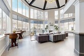Located at 100 Cavalier Crossing in Stonecrest, Georgia, this thoughtfully designed community space makes a striking architectural statement from the moment you enter. The centerpiece is a grand, octagonal great room distinguished by soaring two-story ceilings framed with bold dark beams and expansive floor-to-ceiling windows that wrap the entire perimeter. This dramatic glasswork floods the space with abundant natural light while offering sweeping views of the surrounding community. Light hardwood flooring, structural columns with paneled bases, and a stone-accented fireplace add refined character throughout.

The open layout accommodates multiple seating arrangements, including generous sectional sofas and bar-height dining tables positioned along the windowed walls—ideal for both relaxed gatherings and casual conversations. A mezzanine-level balcony overlooks the main floor, adding vertical depth and architectural interest.

A separate lounge area features rich navy-blue accent walls, crown molding, and large windows that create an intimate yet sophisticated retreat. The kitchen is equally well-appointed, showcasing white shaker cabinetry, dark subway tile backsplash, granite countertops, stainless steel appliances, pendant lighting, and a peninsula with bar seating. Bold navy walls and matte black fixtures give this area a contemporary, polished feel throughout.