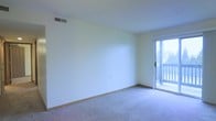 This single-floor space presents a straightforward and functional layout centered around a spacious main living area. The room features neutral, light-colored walls that contribute to a bright and airy atmosphere, complemented by soft carpeted flooring in a muted tone that adds warmth underfoot. A ceiling-mounted HVAC vent ensures climate control throughout the space.

A standout feature of the living area is the sliding glass door positioned along one wall, which floods the interior with natural light and provides direct access to an outdoor balcony. The balcony, partially visible through the glass, extends the living space outdoors and offers additional ventilation options. The door's large glass panels frame exterior views and enhance the overall sense of openness within the room.

The walls are finished cleanly with light-toned paint, and wood-toned baseboards run along the perimeter, providing a subtle contrast to the carpeted floor. The room's open, unobstructed layout offers flexibility for a variety of furniture arrangements. Overall, the space maintains a simple, clean aesthetic suitable for comfortable everyday living.