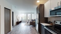 This well-appointed single-floor unit offers a thoughtfully designed layout that balances open communal spaces with private retreats. The open-concept living and dining area is anchored by wide-plank wood-look flooring and bathed in natural light from a bank of large windows with slatted blinds, offering views of surrounding greenery. The space flows seamlessly into a fully equipped kitchen featuring two-tone cabinetry — white upper cabinets paired with dark wood-grain lower cabinets — complemented by light quartz countertops and a full suite of stainless steel appliances, including a refrigerator, range, microwave, and dishwasher. A secondary sitting area, carpeted and quietly tucked away, provides a versatile flex space ideal for a home office or lounge. Both bedrooms are carpeted and filled with natural light from generously sized windows. The primary bedroom includes direct access to an en-suite bathroom, while a second full bathroom is accessible from the main hallway. Both bathrooms are finished with wood-look flooring, white vanity cabinetry, light stone countertops, and walk-in showers. The overall aesthetic is clean, modern, and cohesive, with a neutral palette throughout.