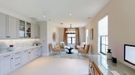 This refined single-floor space blends sophisticated design with functional living. The main lounge area serves as the centerpiece, anchored by a striking circular chandelier with exposed bulbs that casts warm light over a carefully curated seating arrangement of woven accent chairs and a sleek black coffee table. A soft, ocean-toned area rug grounds the space, while an ornate sunburst mirror adds a decorative focal point on the wall. Neutral, warm-toned walls and large windows dressed with flowing drapes allow ample natural light to fill the room, creating an airy and welcoming ambiance throughout.

Adjacent to the lounge, a well-appointed wet bar features gray shaker-style cabinetry with glass-front upper cabinets, a marble mosaic tile backsplash, and a crisp white countertop — ideal for entertaining. A decorative accent wall with textured tile panels and wall sconces adds visual interest to the transitional corridor leading into this area.

The home office is both spacious and practical, accommodating a dual-workstation glass-top desk with built-in drawers and two ergonomic chairs. Multiple windows offer abundant natural light, and the seamless large-format white tile flooring found throughout the space ties all areas together cohesively, reinforcing a clean, modern coastal aesthetic.