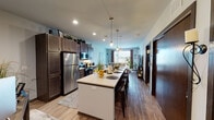 This single-floor residence is designed with an open-concept layout that seamlessly connects the kitchen, dining, and living areas, creating a cohesive and functional living environment. The kitchen is a standout feature, anchored by a generous island with seating and pendant lighting overhead. Rich dark-brown cabinetry extends to the ceiling, providing ample storage, while stainless steel appliances — including a full-size refrigerator, microwave, and range — lend a modern, polished look. Wood-look flooring runs throughout the main living areas, adding warmth and continuity.

The living room is bright and welcoming, benefiting from large windows that invite natural light and offer views of the surrounding neighborhood. A media console with a built-in electric fireplace serves as a focal point, complementing the comfortable, open layout.

The space includes two bedrooms, each carpeted for comfort and featuring windows with exterior views. The primary bedroom offers generous floor space and a relaxed, restful atmosphere. The bathroom is finished with large-format stone-look tile flooring, a dark wood vanity with an integrated sink, and a well-lit mirror, delivering a clean and refined aesthetic throughout.