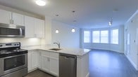 This well-appointed single-floor space showcases a thoughtfully designed open-concept layout that seamlessly connects the kitchen and living areas. The kitchen is a standout feature, outfitted with crisp white shaker-style cabinetry, light granite countertops with subtle veining, and a central island that doubles as a prep and gathering space. A stainless steel undermount sink and dishwasher add a polished, modern touch, while two stylish pendant lights hang above the island, providing focused task lighting. Crown molding along the ceiling adds a refined architectural detail throughout the space. The adjoining living area is bathed in natural light streaming through a large bay window, creating a warm and inviting atmosphere. Dark hardwood-style flooring runs throughout, offering a rich contrast to the bright white walls and cabinetry. Flush ceiling lighting ensures the space remains well-lit at all hours. The open flow between the kitchen and living area makes the overall footprint feel expansive and versatile, ideal for both everyday living and entertaining. The clean, neutral palette and quality finishes give this space a contemporary yet timeless aesthetic.