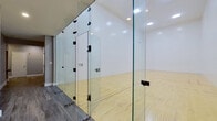 This single-level space is purpose-built around a full-sized racquetball court, offering a well-appointed venue for both competitive and recreational play. The court itself features a light-toned hardwood maple floor with clearly marked red boundary and service lines, providing excellent traction and visibility during play. Tall, smooth white walls surround three sides of the court, meeting the high ceiling overhead, which is equipped with recessed flush-mounted lighting that evenly illuminates the playing surface. A telltale cutout is visible on the front wall, consistent with standard racquetball court specifications.

The court is enclosed on the rear by full-height tempered glass panels with black hardware, allowing clear sightlines into the court from the adjacent viewing corridor. This corridor runs alongside the court and features dark wood-look flooring and neutral grey walls, creating a clean transition between the active play area and surrounding space. Entry to the court is through frameless glass doors. A small anteroom or lobby area is accessible from the corridor, finished with recessed lighting, grey tile wainscoting, and space for posted court rules or signage. The overall design is clean, functional, and purpose-driven.