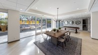 This well-appointed single-story space combines functionality and modern design across several distinct zones. The main gathering area centers around a substantial reclaimed wood dining table set atop a patterned area rug, surrounded by contemporary seating. A statement geometric-patterned accent wall adds visual interest, while large floor-to-ceiling sliding glass doors flood the space with natural light and provide direct access to an outdoor patio featuring tiered seating and landscaping. A billiards table is positioned nearby for recreational use.

The lounge area features a generously sized sectional sofa in deep charcoal, complemented by natural wood slab coffee tables and additional seating. Glass partition walls divide the space into defined areas while maintaining visual openness throughout. Herringbone-patterned light wood flooring runs consistently through the interior zones, contributing to a cohesive aesthetic.

The kitchen is well-equipped with a large central island and bar seating, an oversized range hood, hexagonal tile backsplash, stainless steel appliances, and open shelving. Globe pendant lighting adds warmth. The overall ambiance is polished and contemporary, balancing professional functionality with inviting communal comfort.