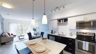 This single-floor 1-bedroom, 1-bathroom residence offers a thoughtfully designed open-concept layout that maximizes every square foot. The kitchen is a standout feature, showcasing a bold two-tone design with sleek dark lower cabinetry contrasted against crisp white upper cabinets and a clean white countertop. A stainless steel dishwasher and undermount sink add modern functionality, while a track lighting system and elegant pendant lights suspended above the kitchen island ensure ample illumination throughout the space. The kitchen island doubles as a dining area, providing a casual yet stylish setting for everyday meals. The open layout flows seamlessly into the living and dining areas, where natural light pours in through large windows that overlook a lush, green outdoor setting, creating a bright and airy ambiance. The living area offers a comfortable and inviting retreat, with a neutral color palette and tasteful décor that complement the home's contemporary aesthetic. The combination of hardwood-style flooring, high ceilings, and generous natural light enhances the overall sense of openness. This residence strikes an ideal balance between modern design and practical living.