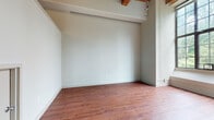 This single-floor space showcases a distinctive loft-inspired design that blends industrial character with residential functionality. Throughout the main living areas, warm-toned hardwood flooring runs continuously, complementing the exposed wood beam ceilings overhead. The structural beams add architectural depth and a raw, industrial aesthetic that defines the overall ambiance of the space.

The open living area benefits from generous ceiling height, with large multi-pane industrial-style windows allowing substantial natural light to filter in, offering views of surrounding greenery. A staircase is visible leading to an upper mezzanine or storage level, adding vertical dimension to the layout. Track lighting along the beams provides additional illumination.

The kitchen is efficiently laid out in an L-shaped configuration, featuring warm honey-toned wood cabinetry with brushed silver hardware, white countertops, a stainless steel sink, a tile backsplash, and under-cabinet lighting. A dishwasher and range are included among the appliances.

The bathroom is cleanly finished with white tile walls, a tub-shower combination, a white vanity with a single basin sink, and a large mirror, creating a bright and functional space. Overall, the unit presents a cohesive industrial-residential character throughout.