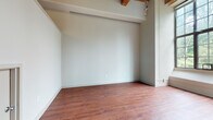 This single-floor space showcases a distinctive loft-inspired design that blends industrial character with residential functionality. Throughout the main living areas, warm-toned hardwood flooring runs continuously, complementing the exposed wood beam ceilings overhead. The structural beams add architectural depth and a raw, industrial aesthetic that defines the overall ambiance of the space.

The open living area benefits from generous ceiling height, with large multi-pane industrial-style windows allowing substantial natural light to filter in, offering views of surrounding greenery. A staircase is visible leading to an upper mezzanine or storage level, adding vertical dimension to the layout. Track lighting along the beams provides additional illumination.

The kitchen is efficiently laid out in an L-shaped configuration, featuring warm honey-toned wood cabinetry with brushed silver hardware, white countertops, a stainless steel sink, a tile backsplash, and under-cabinet lighting. A dishwasher and range are included among the appliances.

The bathroom is cleanly finished with white tile walls, a tub-shower combination, a white vanity with a single basin sink, and a large mirror, creating a bright and functional space. Overall, the unit presents a cohesive industrial-residential character throughout.