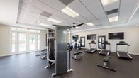 This fitness center is a comprehensively equipped, single-floor facility designed to support a full range of workout routines. The space is anchored by a suspended tile ceiling fitted with large panel LED lighting and an oversized ceiling fan, ensuring the room remains bright and well-ventilated throughout. Durable rubber flooring covers the entire area, providing traction and impact absorption across all workout zones.

The cardio section is well-stocked with multiple treadmills, elliptical trainers, upright and recumbent stationary bikes, and a stair climber. A multi-station cable machine anchors the strength training area, complemented by a free weight rack stocked with dumbbells, a stability ball, and adjustable benches for varied exercises. Wall-mounted flat-screen televisions and full-length mirrors in dark frames are positioned throughout, enhancing both entertainment and form-checking during workouts.

A bold royal blue accent wall adds a dynamic visual element to the free weights corner, contrasting with the neutral tones of the surrounding walls. Multiple sets of glass-paneled French doors with transom windows line one side of the room, flooding the space with natural light and offering direct views of and access to an outdoor pool and patio area. The overall design is clean, functional, and energizing.