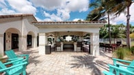 This single-story outdoor amenity space showcases a beautifully designed Mediterranean-inspired cabana that serves as the centerpiece of a resort-style recreational area. The structure features a distinctive terracotta tile roof, sturdy stucco-finished columns with arched openings, and a ceiling fan overhead, creating a comfortable, shaded gathering space even on warm sunny days. At the heart of the cabana sits a generously sized built-in bar with a stone or tile-finished counter, surrounded by tall bar-height chairs, making it an ideal space for socializing and entertaining. The surrounding patio is finished with large-format, neutral-toned pavers that lend a clean, upscale aesthetic to the outdoor environment. Lush tropical landscaping, including mature palm trees, frames the space and reinforces the resort-like atmosphere. In the background, a pool area with lounge seating and market umbrellas is visible, suggesting a cohesive and well-appointed outdoor amenity complex. The open-air design allows for abundant natural light and refreshing breezes, while the covered structure provides shade and shelter. The overall ambiance is relaxed yet refined, evoking the feel of a Florida or Caribbean retreat suited for leisure and community gatherings alike.