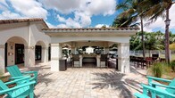 This single-story outdoor amenity space showcases a beautifully designed Mediterranean-inspired cabana that serves as the centerpiece of a resort-style recreational area. The structure features a distinctive terracotta tile roof, sturdy stucco-finished columns with arched openings, and a ceiling fan overhead, creating a comfortable, shaded gathering space even on warm sunny days. At the heart of the cabana sits a generously sized built-in bar with a stone or tile-finished counter, surrounded by tall bar-height chairs, making it an ideal space for socializing and entertaining. The surrounding patio is finished with large-format, neutral-toned pavers that lend a clean, upscale aesthetic to the outdoor environment. Lush tropical landscaping, including mature palm trees, frames the space and reinforces the resort-like atmosphere. In the background, a pool area with lounge seating and market umbrellas is visible, suggesting a cohesive and well-appointed outdoor amenity complex. The open-air design allows for abundant natural light and refreshing breezes, while the covered structure provides shade and shelter. The overall ambiance is relaxed yet refined, evoking the feel of a Florida or Caribbean retreat suited for leisure and community gatherings alike.