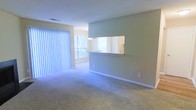 This single-floor space offers a straightforward, functional layout designed for comfortable everyday living. The living area is finished with wall-to-wall carpeting in a neutral tone, providing a soft underfoot feel throughout. A large window dressed with vertical blinds allows natural light to filter into the space, creating a bright and airy atmosphere while offering privacy control.

A defining architectural feature is the pass-through opening in the dividing wall between the living area and the kitchen. This cut-out enhances the sense of openness and connectivity between the two spaces, allowing light to travel freely and making the overall layout feel more expansive. The kitchen, visible through the pass-through, includes overhead lighting and appears to benefit from its own window, adding to the natural brightness of the unit.

The walls are finished in a soft, neutral tone that complements the light carpeting and contributes to an understated, calm ambiance. White baseboards provide clean visual definition along the floor line. The space is currently vacant, allowing the layout and architectural details to be clearly appreciated.