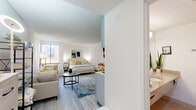 Located at 2444 E Maple Ave in Burlington, NC, this studio unit is thoughtfully designed to maximize every square foot of its single-floor layout. The open-plan main area seamlessly integrates sleeping, living, and dining functions into one bright, cohesive space. Light gray luxury vinyl plank flooring runs throughout, complementing the crisp white walls and textured ceiling that lend a clean, modern aesthetic.

A large window along the exterior wall floods the living area with natural light, creating an airy atmosphere. The sleeping zone accommodates a full-sized bed and nightstands, while a compact dining set with two chairs provides a dedicated eating area. A wall-mounted television adds convenience and function without sacrificing floor space.

The kitchenette is efficiently outfitted with gray shaker-style cabinetry, a smooth cooktop, single-basin sink, upper storage cabinets, and a full-sized stainless steel refrigerator with a microwave above. This compact culinary area is situated to keep the main living space open and unobstructed.

The bathroom features tile flooring, a wide granite-topped vanity with an under-mount sink, a large mirror, and ample towel storage. Overall, the unit offers a well-organized, functional layout suited for single occupancy in a convenient Burlington location.