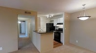 This single-floor residential unit offers a practical and well-organized layout that maximizes everyday livability. The heart of the home is an open-concept kitchen and living area, where a peninsula-style breakfast bar with a dark granite countertop serves as a natural divider between the two spaces. The kitchen is equipped with black appliances, including a refrigerator, electric range, and over-the-range microwave, all complemented by crisp white cabinetry that provides ample storage. A track lighting fixture overhead ensures the kitchen is well-lit. The flooring transitions from warm hardwood in the kitchen to plush carpeting in the living area, adding a sense of comfort underfoot. Neutral, warm-toned walls run throughout the space, creating a cohesive and welcoming ambiance. A hallway visible from the main living area leads to additional rooms, suggesting a thoughtfully planned floor plan with private spaces set apart from the main living and dining zones. Ventilation is provided through ceiling vents, and the overall layout reflects a clean, functional design suited for comfortable day-to-day living.