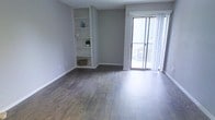 This single-floor space offers a clean, modern aesthetic designed for comfortable everyday living. The main living area is finished with rich, dark hardwood-style plank flooring that runs throughout, adding warmth and a sense of continuity to the space. Neutral grey walls create a calm, versatile backdrop that complements a wide range of furnishing styles.

A standout architectural feature is the built-in wall shelving unit, thoughtfully recessed into the wall to provide display and storage space without encroaching on the living area. The shelves offer a practical yet stylish element, ideal for books, décor, or personal items.

Natural light flows generously into the room through a full-height sliding glass door fitted with sheer vertical blinds, which allow light to be filtered while maintaining privacy. The sliding door opens to an outdoor area, providing a seamless connection between interior and exterior living spaces.

The overall layout is open and uncluttered, offering flexibility in how the space can be arranged and utilized. Clean baseboards and well-maintained finishes throughout speak to the quality and upkeep of the space. The combination of functional built-ins, abundant natural light, and contemporary flooring makes this a welcoming and practical living environment.