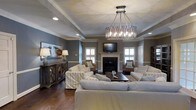This single-floor residence showcases a thoughtfully designed living space that balances classic elegance with relaxed, coastal-inspired comfort. The living room serves as the heart of the home, drawing the eye upward to a tray ceiling adorned with crisp crown molding and anchored by a striking drum-style chandelier with an ornate metal frame and candelabra lighting. Recessed lighting complements the chandelier, ensuring the room is well-lit throughout. Walls finished in a warm, muted gray tone create a sophisticated yet inviting atmosphere, harmonizing beautifully with the rich dark hardwood flooring that runs throughout the space. A white-mantled fireplace serves as a natural focal point, flanked by shuttered windows that allow for controlled natural light. A wall-mounted television above the fireplace adds a modern, functional touch. The seating arrangement is generous and welcoming, featuring plush sofas and striped armchairs that evoke a relaxed, coastal sensibility. A rustic wooden console table along one wall provides both storage and decorative display space, complemented by coordinating table lamps. The overall ambiance is one of refined casualness — a space designed for both comfortable everyday living and gracious entertaining.