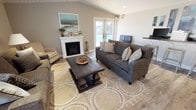 Located at 3000 Austin Dr in Bremerton, Washington, this single-story home offers a thoughtfully designed interior with a welcoming and comfortable atmosphere throughout. The living room serves as the heart of the home, showcasing vaulted ceilings with white shiplap paneling and a ceiling fan that enhance the sense of openness. A white-mantled fireplace anchors one wall, while French doors on the opposite side allow natural light to pour in and provide views of the outdoor area. Wood-look flooring runs throughout, complementing the neutral warm-toned walls. Seating is arranged around a bold dark wood coffee table atop a patterned area rug, creating a relaxed and inviting gathering space. Adjacent to the living area, a built-in counter with bar seating offers a functional workspace or casual dining spot. The kitchen features white shaker-style cabinetry paired with stainless steel appliances, including a refrigerator, range, microwave, and dishwasher, all set against dark countertops. A separate dining area accommodates a four-person table and benefits from abundant natural light through a large window with plantation-style blinds, making it an ideal spot for everyday meals.