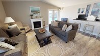 Located at 3000 Austin Dr in Bremerton, Washington, this single-story home offers a thoughtfully designed interior with a welcoming and comfortable atmosphere throughout. The living room serves as the heart of the home, showcasing vaulted ceilings with white shiplap paneling and a ceiling fan that enhance the sense of openness. A white-mantled fireplace anchors one wall, while French doors on the opposite side allow natural light to pour in and provide views of the outdoor area. Wood-look flooring runs throughout, complementing the neutral warm-toned walls. Seating is arranged around a bold dark wood coffee table atop a patterned area rug, creating a relaxed and inviting gathering space. Adjacent to the living area, a built-in counter with bar seating offers a functional workspace or casual dining spot. The kitchen features white shaker-style cabinetry paired with stainless steel appliances, including a refrigerator, range, microwave, and dishwasher, all set against dark countertops. A separate dining area accommodates a four-person table and benefits from abundant natural light through a large window with plantation-style blinds, making it an ideal spot for everyday meals.
