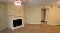 This single-floor space presents a comfortable and straightforward layout centered around a welcoming living area. The room is anchored by a classic fireplace framed with a white mantel and a tiled hearth, serving as the focal point of the space. Warm beige walls and wall-to-wall carpeting in a neutral tan tone create a cohesive, understated aesthetic throughout. A decorative ceiling light fixture adds a subtle ornamental touch overhead, while a ceiling vent ensures consistent climate control. The open wall space adjacent to the fireplace offers flexibility for furniture arrangement and personalization. A doorway visible toward the back of the room provides access to additional areas of the unit, suggesting a practical and connected floor plan. The textured ceiling is consistent with the overall construction style. The space is well-suited for comfortable everyday living, with its neutral palette providing a versatile backdrop for a variety of interior design preferences. Overall, the space conveys a lived-in warmth with functional simplicity.