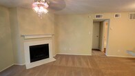 This single-floor space presents a comfortable and straightforward layout centered around a welcoming living area. The room is anchored by a classic fireplace framed with a white mantel and a tiled hearth, serving as the focal point of the space. Warm beige walls and wall-to-wall carpeting in a neutral tan tone create a cohesive, understated aesthetic throughout. A decorative ceiling light fixture adds a subtle ornamental touch overhead, while a ceiling vent ensures consistent climate control. The open wall space adjacent to the fireplace offers flexibility for furniture arrangement and personalization. A doorway visible toward the back of the room provides access to additional areas of the unit, suggesting a practical and connected floor plan. The textured ceiling is consistent with the overall construction style. The space is well-suited for comfortable everyday living, with its neutral palette providing a versatile backdrop for a variety of interior design preferences. Overall, the space conveys a lived-in warmth with functional simplicity.