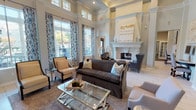 This single-floor communal space exemplifies refined design through its dramatic, soaring ceilings adorned with detailed crown molding and a coffered ceiling treatment that adds architectural depth. Tall, floor-to-ceiling windows dressed in bold blue-and-white ikat drapery flood the main lounge area with generous natural light while offering outdoor views. The centerpiece of the lounge is a stately white-stone fireplace with an ornate mantel, flanked by a wingback chair and complemented by a curated arrangement of a dark upholstered sofa, patterned area rug, and a sleek glass-and-chrome coffee table. Structural columns with classical detailing define distinct zones throughout the open layout. A built-in shelving unit in a slate-blue finish houses a mounted television and display accents, while a compact desk station with a marble-topped table and ornate carved chair provides a functional nook. Artwork in neutral tones decorates the walls, contributing to the sophisticated yet welcoming ambiance. Light-toned tile flooring runs throughout, enhancing the bright, airy atmosphere. The space transitions seamlessly between the lounge, a secondary seating area, and what appears to be a business or service counter, reflecting a thoughtfully designed, multi-functional environment.