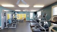 Located at 405 Grand Wailea Dr in Fayetteville, NC, this fitness center is a well-appointed, single-floor facility designed to support a complete workout experience. The main exercise area features teal-painted walls that create an energizing atmosphere, complemented by durable dark rubber flooring ideal for high-traffic gym use. Overhead, dual ceiling fans work in tandem with recessed fluorescent lighting to maintain a comfortable and well-lit environment throughout the space. The cardio zone is lined with a row of Star Trac treadmills and elliptical machines positioned alongside windows that allow natural light to filter in, offering a refreshing view while exercising. On the opposing side, a multi-station strength training unit and an adjustable weight bench provide options for resistance and functional training. Wall-mounted televisions are positioned for visibility from both the cardio and strength training areas, enhancing the workout experience. An open doorway leads to an adjoining room with bright yellow walls, which appears to serve as a separate activity or lounge area with its own distinct character. This fitness center is a thoughtfully organized space that balances functionality with a welcoming, vibrant aesthetic.
