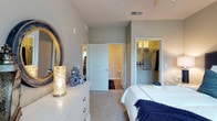 This single-floor residence offers a thoughtfully designed layout with a refined sense of comfort and practicality. The bedroom serves as a serene retreat, finished with soft neutral tones on the walls and plush carpeted flooring that adds warmth underfoot. A white dresser with classic hardware provides generous storage and is complemented by decorative accents, including blue-and-white ceramic vases and a sculptural accent lamp, creating an elegant yet welcoming atmosphere. An ornate gold-framed mirror adds a touch of sophistication to the space. The bedroom opens conveniently to both an ensuite bathroom and a spacious walk-in closet, offering excellent organization and privacy. The walk-in closet is equipped with built-in shelving and hanging rods, providing ample room for wardrobe storage. Neutral gray walls and clean white trim carry throughout the space, lending a cohesive and contemporary aesthetic. Overhead ventilation and recessed lighting ensure year-round comfort, while the open doorway to the adjoining living area hints at a well-connected and functional floor plan. Overall, this residence balances practical amenities with understated elegance.