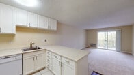 This single-floor residence offers a practical and functional layout with a clean, neutral aesthetic throughout. The kitchen is well-appointed with white cabinetry, light speckled countertops, a stainless steel sink, and tile flooring. A peninsula counter extends into the living area, creating an open-concept flow and additional workspace. White appliances—including a refrigerator, oven, and dishwasher—complete the kitchen's practical design. The living area is generously sized, featuring wall-to-wall carpeting, warm beige walls, and a textured ceiling. A sliding glass door leads to an outdoor balcony, allowing natural light to fill the space and providing an outdoor view. A wall-mounted HVAC unit ensures climate control convenience. The bedroom is spacious and carpeted, featuring a large window with vertical blinds that invites natural light and a wall-mounted HVAC unit. The room's open layout offers flexibility in furniture arrangement. The bathroom is finished with tile flooring and includes a full-size tub and shower combination enclosed by sliding glass doors with chrome hardware. Throughout the space, the consistent use of light, neutral tones creates a cohesive and bright atmosphere.
